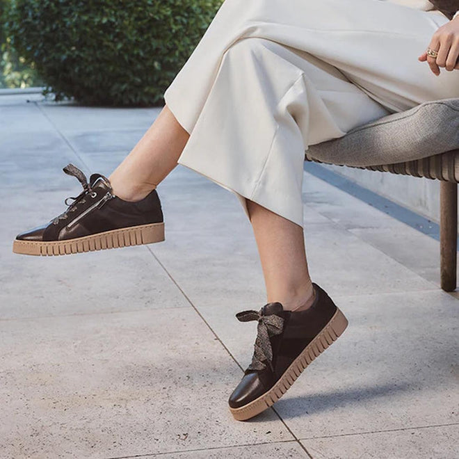 A person sits outdoors on a chair, one foot slightly raised, wearing wide-legged white pants and EOS Tinah Mahogany Dark Brown Leather Trainers With Zip., 51468, 9321190042108, 9321190042115, 9321190042122, 9321190042139, 9321190042146, 51474