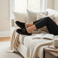 Relaxing on a cozy sofa with neutral pillows and a knitted blanket, someone wears Emu Womens Adison Black Sheepskin Wool Mule Slipper Scuffs. An open book is on their lap, and a coffee table sits nearby., 50710, 9330071990797, 9330071990803, 9330071990810, 50714, 50715