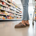 A person wearing Emu Womens Adison Smoke Grey Wool Mule Slippers and blue jeans stands beside a shopping cart in a grocery store aisle, with shelves of various products visible in the background., 50716, 9330071986417, 9330071986424, 9330071986431, 50720, 50721