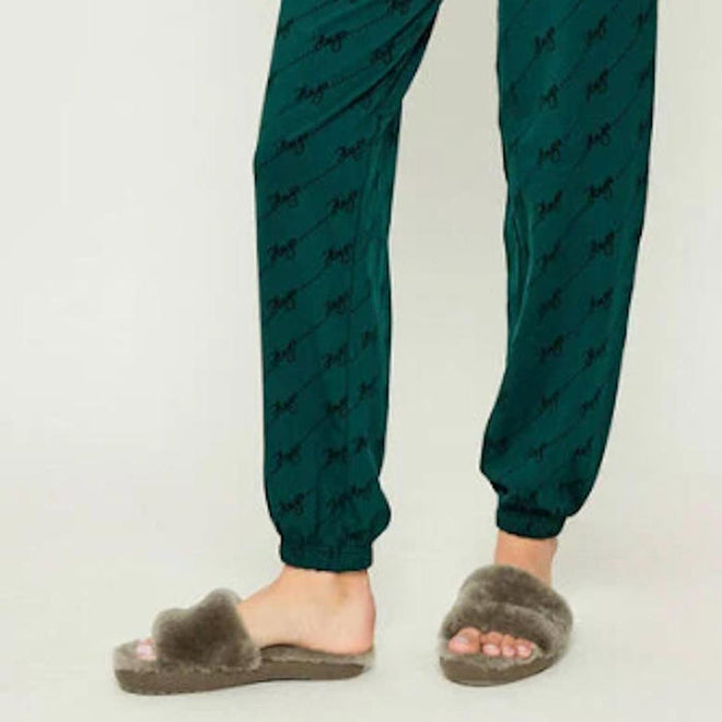 A person wearing green pajama pants stands on a light-colored floor, with only their lower legs and feet visible. They are wearing EMU Women's Mckay Dark Olive Sheepskin Scuff Slipper Slides., 50733, 9330071976265, 9330071976272, 9330071976289, 50737, 50738