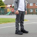 A child in a grey school uniform wears Harrison Nifty Black School Trainers with self-fastening straps, standing on a playground with blurred equipment and houses in the background. The child's face is not visible., 48569, 48570, 48571, 9361913034997, 48573, 9361913035031, 48575, 48576, 48577, 48578, 48579, 48569, 48570, 48571, 9361913034997, 48573, 9361913035031, 9361913035055, 9361913035079, 48577, 9361913035116, 48579