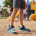 A child in Harrison Nifty Kids Sports Trainer Navy/Green/White shoes with adjustable fit stands on wood chips at a playground, while other children play on swings and a yellow slide in the background., 49664, 9361913035710, 9361913035734, 9361913035758, 9361913035772, 9361913035796, 9361913035819, 9361913035833, 49672, 49673, 49674
