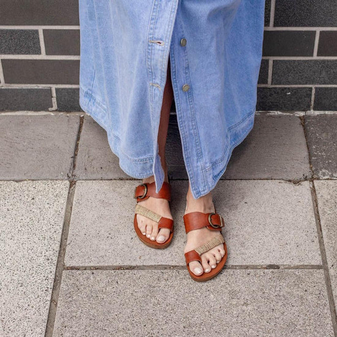A person wearing a light blue buttoned denim skirt and Los Cabos Bria Cognac Light Flexible Sandal Jandals stands on a tiled pavement near a brick wall., 46914, 9321191210063, 9321191210070, 9321191210087, 9321191210094, 9321191210100, 9321191210117