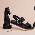 The Los Cabos Sana Black Sandal With Back Strap, a comfortable summer sandal with an adjustable slingback and gold buckle, is displayed beside a large black chess knight on a plain light beige background. Two small gold accessories are positioned on the beige surface in front of them., 9321191314006, 9321191314013, 9321191314020, 9321191314037, 9321191314044, 9321191314051, 53748