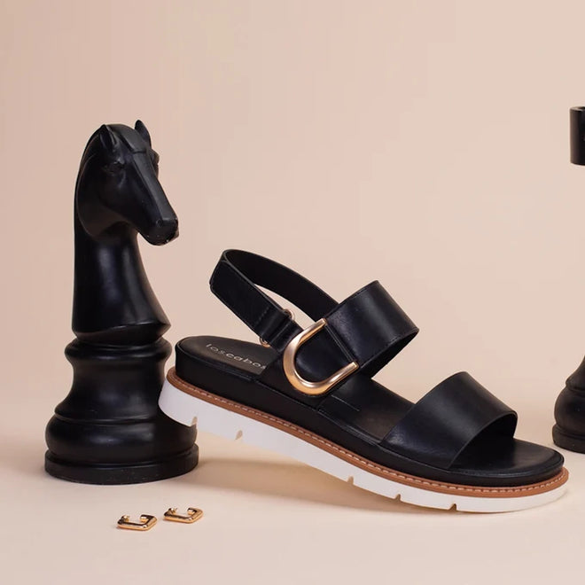 The Los Cabos Sana Black Sandal With Back Strap, a comfortable summer sandal with an adjustable slingback and gold buckle, is displayed beside a large black chess knight on a plain light beige background. Two small gold accessories are positioned on the beige surface in front of them., 9321191314006, 9321191314013, 9321191314020, 9321191314037, 9321191314044, 9321191314051, 53748