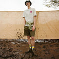 A person stands smiling on muddy ground, wearing a bucket hat, sunglasses, a beige t-shirt with a pink smiley logo, green shorts, patterned socks, Barbour Nimbus Olive/Ochre Chelsea Wellingtons Men's Rainboots, and a sweatshirt tied around their waist., 5063669168813, 5063669168820, 5063669168769, 5063669168776