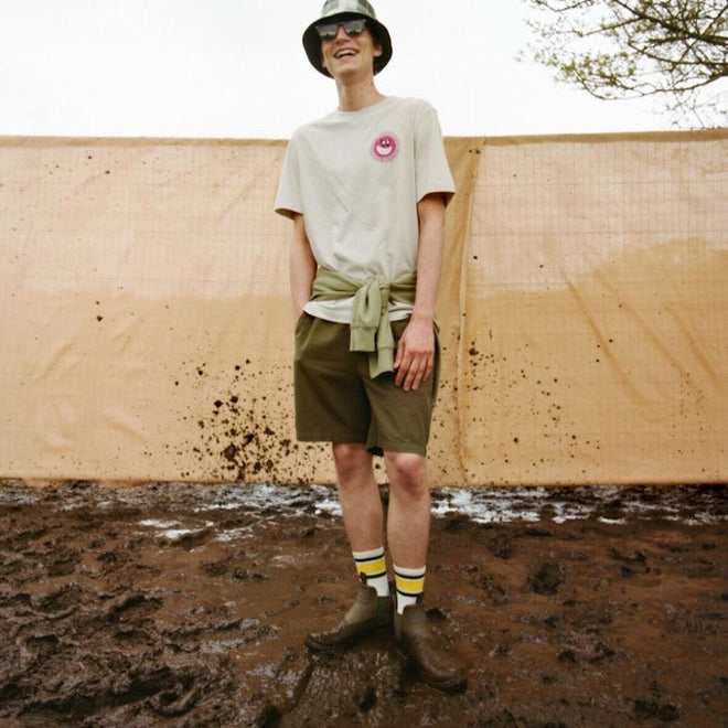 A person stands smiling on muddy ground, wearing a bucket hat, sunglasses, a beige t-shirt with a pink smiley logo, green shorts, patterned socks, Barbour Nimbus Olive/Ochre Chelsea Wellingtons Men's Rainboots, and a sweatshirt tied around their waist., 5063669168813, 5063669168820, 5063669168769, 5063669168776