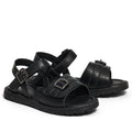 The Pretty Brave Phoebe Girls Black Leather School Sandal features black leather, crisscross straps, open toes, ankle straps, and adjustable metal buckles, shown against a white background.