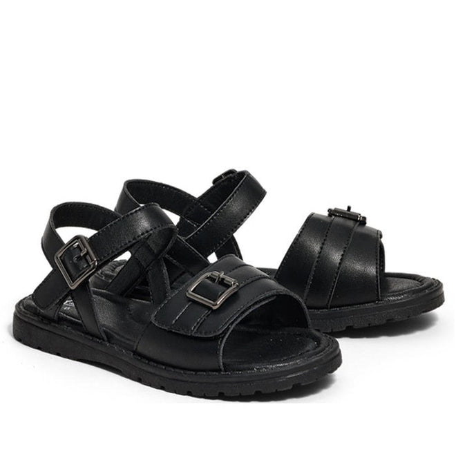 The Pretty Brave Phoebe Girls Black Leather School Sandal features black leather, crisscross straps, open toes, ankle straps, and adjustable metal buckles, shown against a white background.