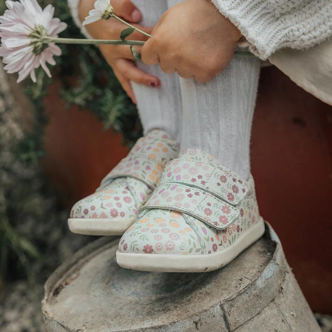Children's floral shoes worn by a child sitting on a stone block with a blurred background.
9420075347700,