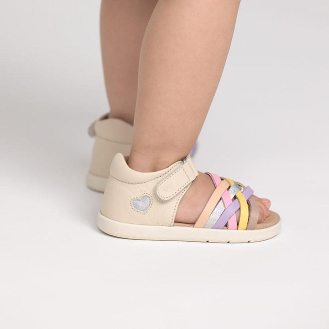 A toddler stands on a white surface wearing Pretty Brave 1st Walker Eden Rainbow Leather Open Toe Sandals, featuring podiatrist-approved design, pastel pink, yellow, purple, and silver straps, a blue heart detail, and an adjustable strap., 9420075355705, 9420075355699, 9420075355682, 9420075355675