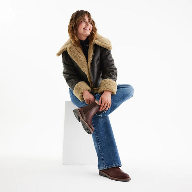 A person wearing a shearling-lined black leather jacket, blue jeans, and Rollie Womens Chelsea Rise Vintage Mocha Leather Boots sits on a white block against a plain white background, smiling with one leg crossed over the other., 9339554341682, 9339554341699, 9339554341705, 9339554341712, 9339554341729