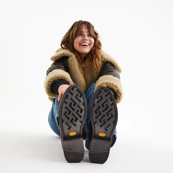 A smiling woman in a shearling jacket and jeans sits on the floor, legs extended to show the Vibram outsole of her Rollie Womens Chelsea Rise Vintage Mocha Leather Boots, against a plain white background., 9339554341682, 9339554341699, 9339554341705, 9339554341712, 9339554341729