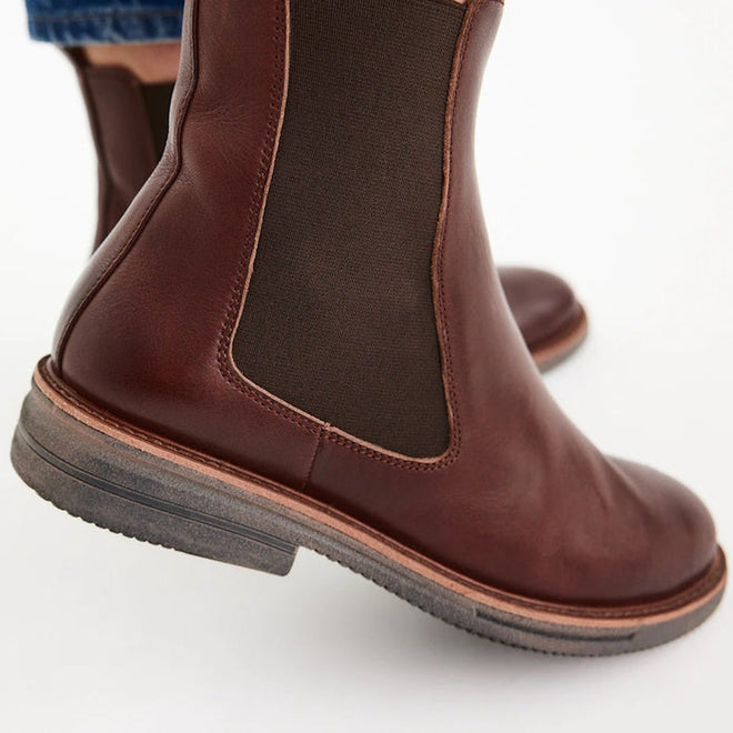 Close-up of a person wearing Rollie Womens Chelsea Rise Vintage Mocha Leather Boot with dark elastic side panels, light brown soles, and a Vibram outsole, standing on a white background., 9339554341682, 9339554341699, 9339554341705, 9339554341712, 9339554341729