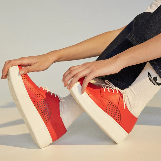 A person in dark blue jeans and white socks with a black Adidas logo sits on the floor, slightly bent forward, touching their Rollie Womens Derby City Punch Red Leather Trainers with white soles., 9781760775599, 9781760775605, 9781760775612, 9781760775629, 9781760775636