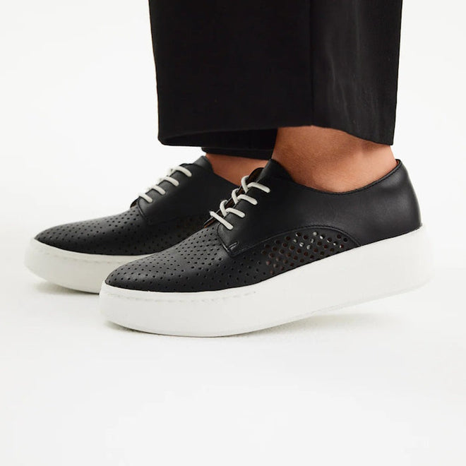 A person wearing Rollie Womens Derby City Punch Black Leather Trainers with thick white platform soles and black pants stands on a white background. These sneakers are orthotic-friendly with a removable footbed for comfort., 9339554259628, 9339554259635, 9339554259642, 9339554259659, 9339554259666, 9339554259673