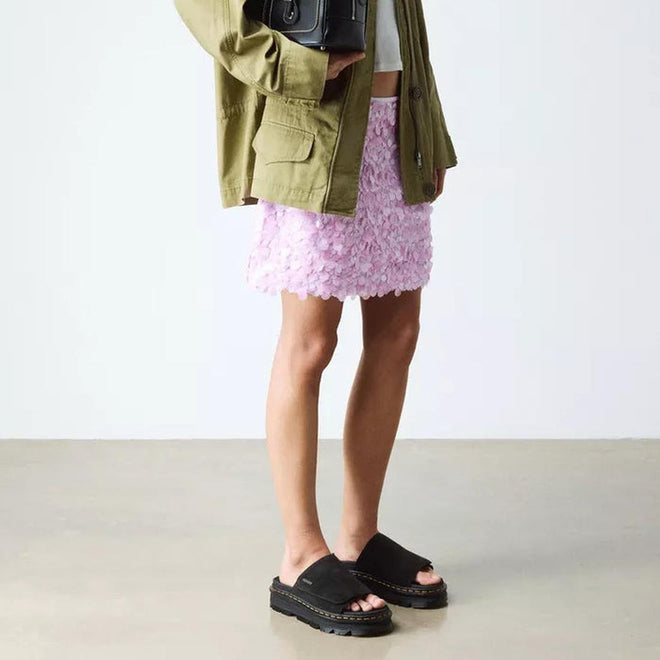 Person wearing an olive green jacket, pale pink textured mini skirt, and Dr Martens Zebzag Black EH Suede Slide Sandals with air-cushioned sole, standing indoors on a light floor against a white background., 190665790689, 190665790696, 190665790702, 190665790726, 190665790733, 52118