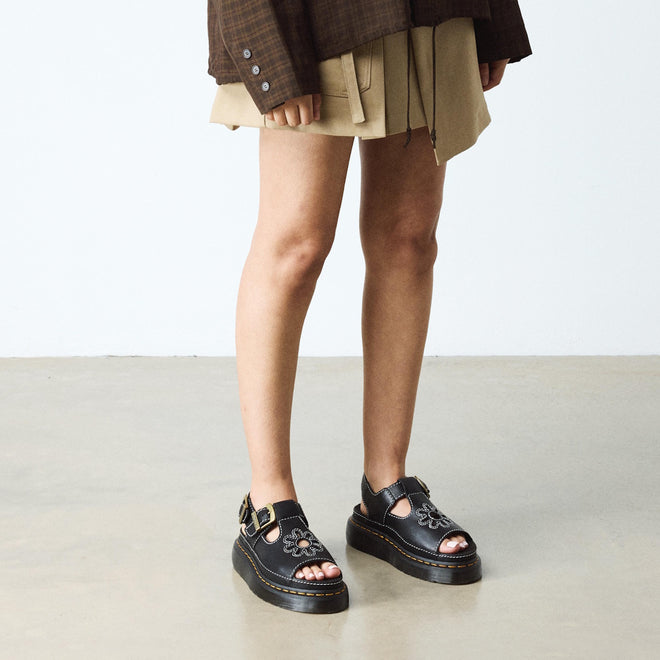 A person wearing a brown plaid shirt, tan skirt, and Dr Martens Dunnet Flower Sandal Black Quad Platform stands on a light-colored floor against a plain white wall., 190665809190, 190665809206, 190665809213, 190665809237, 190665809244, 52092
