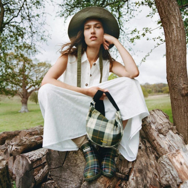 A woman in a white dress and wide-brimmed hat sits on a large log outdoors, holding a plaid bag and wearing Barbour Wilton Classic Tartan Chelsea Wellingtons Womens Rainboot, with trees and grass in the background., 194972145934, 194972146030, 194972146122, 194972146221, 194972146283, 51397