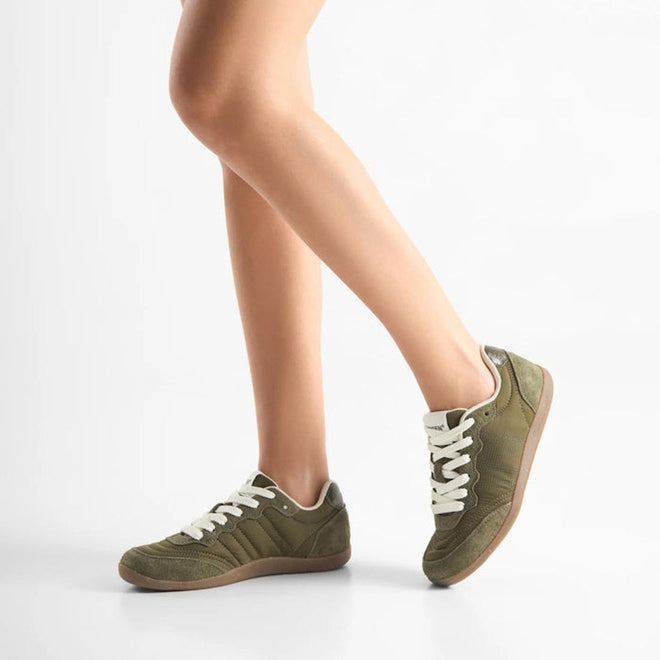 A person stands against a white background, one leg slightly bent, wearing Woden Nanna Dark Olive Lace Up Sneakers with white laces, gum soles, and WODEN Natural Soft Technology., 5713326697506, 5713326697513, 5713326697520, 5713326697537, 5713326697544, 5713326697551