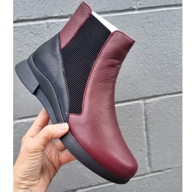 A hand holds up the Alfie & Evie Junior Bordo Purple Lo Leather Ankle Boot Womens, featuring a Bordo purple leather front, black elastic side panel, and memory foam insole, against a gray cinder block wall., 50876, 50877, 50878, 50879, 50880, 50881, 50882