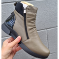 A hand holds the Alfie & Evie Justice Khaki Lo Leather Zip Ankle Boot for women, featuring khaki leather, a side zipper, black patent heel detail, black sole, and removable memory foam insole against a gray textured wall., 50897, 50898, 50899, 50900, 50901, 50902, 50903