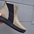 A hand holds the Alfie & Evie Womens Eagle Khaki Chelsea Ankle Boot Zip, featuring a black elastic side panel, thick black rubber sole, and cushioned memory foam insole, against a gray cinder block wall background., 50834, 50835, 50836, 50837, 50838, 50839, 50840