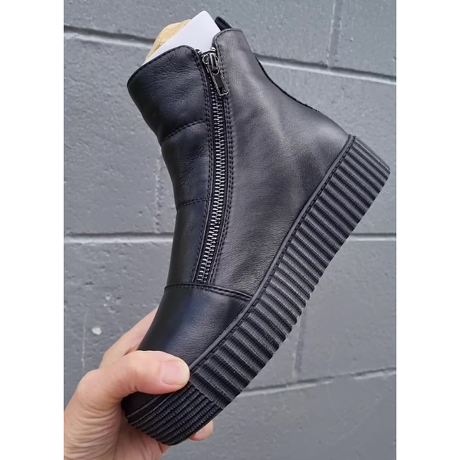 A hand holds the Alfie & Evie Womens Dynasty Black Leather Ankle Boot, featuring a side zipper, thick ribbed creeper sole, and memory innersole, with a gray cinder block wall in the background., 50918, 50919, 50920, 50921, 50922, 50923, 50924