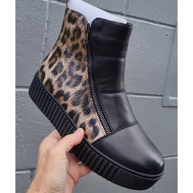 A hand holds the Alfie & Evie Womens Dynasty Black Leopard Cracked Leather Ankle Boot, featuring a thick black platform sole, black leather front, leopard print side panel, and a visible side zipper against a gray brick wall background.