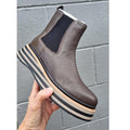 A hand holds up the Alfie & Evie Molten Choc Brown Leather Ankle Boot for women, featuring black elastic side panels, a chunky striped platform sole, set against a gray cinder block wall., 50862, 50863, 50864, 50865, 50866, 50867, 50868
