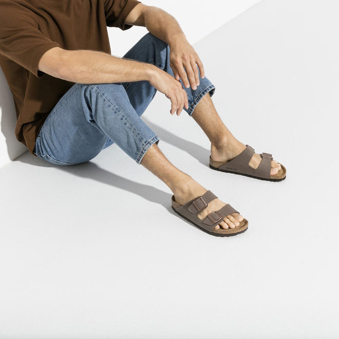 A man sittimg down against a wall showing a pair of mocca birkenstocks on the feet. knees bent.  4040714059273,32651,32653,4040714059280,4040714059297,4040714059303,4040714059310,4040714059327,4040714059334,,,,

