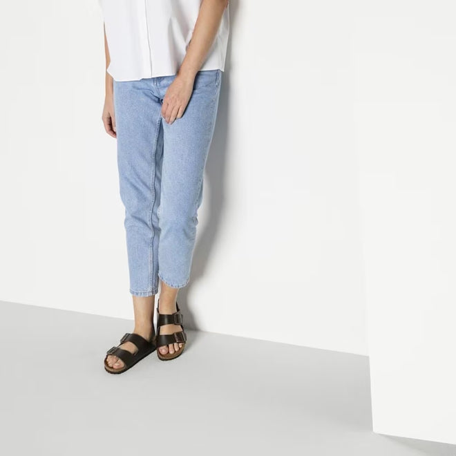 A modela wearing light blue denim 3/4 pants and a white shirt standing against a wall wearing Birkenstock Arizona leather sandal slides in dark bown narrow fit. showing double straps across the top of the foot.,4013871078616,4013871018407,4013871019466,4013871019558,4013871045779,4013871058250,4013871059127,4013871074724, , , , , 
