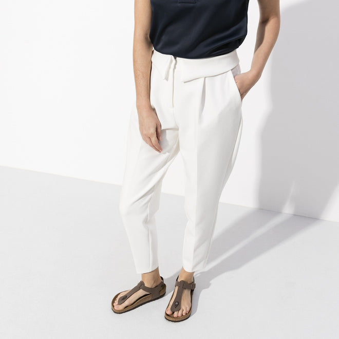 A person wearing a sleeveless black top, high-waisted white trousers, and BIRKENSTOCK Kairo Womens Birkibuc Mocha Brown Regular Width sandals stands against a white background with one hand in their pocket. Their face is not visible., 4052001079413, 4052001079420, 4052001079437, 4052001079444, 4052001079451, 50471, 50472