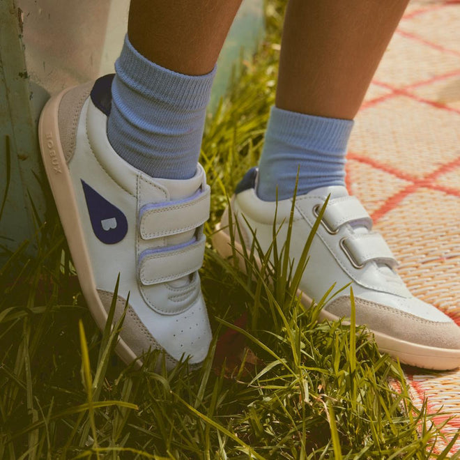 Close-up of a person wearing light blue socks and Bobux Kids IWalk IW Champ Sneaker Chalk White Blue 650501, featuring velcro straps and a blue side design, standing on green grass near a patterned outdoor mat., 9420075767843, 9420075781443, 9420075780316, 9420075781368, 9420075780460
