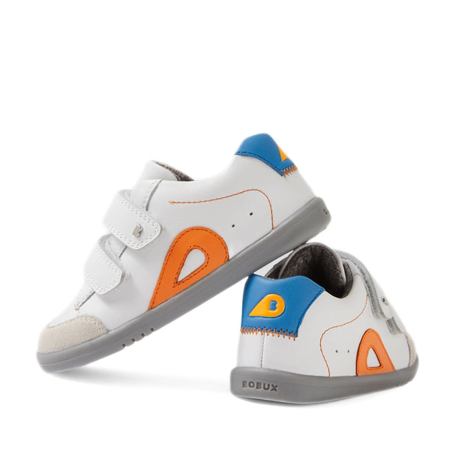 Two Bobux IW Iwalk Blaze Toddlers Leather Sneakers in White Orange, featuring gray soles, orange and yellow side accents, blue heel tabs, and Velcro straps. Crafted from quick-drying leather, one sneaker stands upright while the other is tilted to display the back and side.