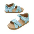 A pair of Bobux IWalk Flora Jelly Mint Metallic Blue open toe sandals for toddlers, featuring a tan sole, Velcro straps, and a subtle star and swirl pattern.