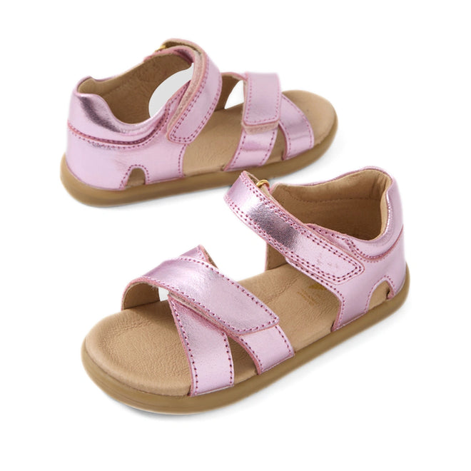 The Bobux IWalk Flora Toddlers Sandal in Pink Lemonade is a pair of metallic pink children’s sandals with crisscross straps, open toes, ankle support, and secure velcro closures. Made from breathable leather with tan soles and a cushioned insole, these sandals ensure all-day comfort for little feet.
