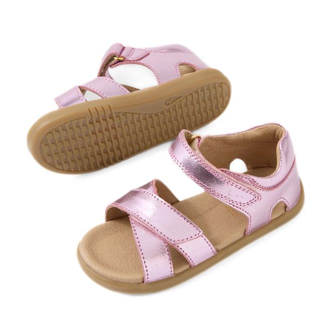 Introducing the Bobux IWalk Flora Toddlers Sandal in Pink Lemonade: a pair of metallic pink open-toe sandals for children, featuring breathable leather uppers, tan rubber soles, and adjustable hook-and-loop straps for a secure fit. Perfectly crafted for toddlers, one sandal is shown from the side while the other displays its sole.