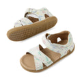 Two white Bobux IWalk Flora Toddlers Sandals featuring a pastel animal and floral pattern, tan soles, open toes, and adjustable Velcro ankle straps. One sandal is shown from the top view while the other displays the bottom sole. These leather toddler sandals perfectly combine stylish comfort for little feet on every adventure.