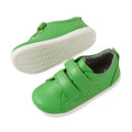 A pair of Bobux Iwalk Toddlers Grass Court Retro Green Leather Trainers, featuring white soles, Velcro straps, and small ventilation holes on the side. One shoe is lying flat while the other is on its side showcasing the sole—perfect toddler trainers for everyday adventures., 9420074806789, 9420074806796, 9420074806802, 9420074806819, 9420074806826