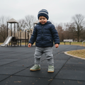 A toddler in a blue jacket, gray knit pants, and striped beanie stands on a playground wearing Bobux IW Iwalk Maverick Leather Sneaker Vetiver Green. The overcast sky and leafless trees complete the scene., 9420074836427, 9420074836434, 9420074836441, 9420074836458, 9420074836465