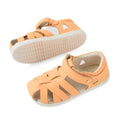 A pair of Bobux Iwalk IW Tropicana II Peach Jelly Leather Sandals, crafted from peach-colored leather with woven straps, ankle buckles, and durable white rubber soles that provide closed-toe protection. One sandal is displayed sole-up while the other rests flat., 9420074810083, 9420074810090, 9420074810106, 9420074810113, 9420074810120