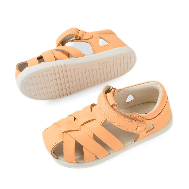 A pair of Bobux Iwalk IW Tropicana II Peach Jelly Leather Sandals, crafted from peach-colored leather with woven straps, ankle buckles, and durable white rubber soles that provide closed-toe protection. One sandal is displayed sole-up while the other rests flat., 9420074810083, 9420074810090, 9420074810106, 9420074810113, 9420074810120