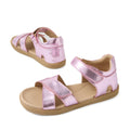 Two Bobux KP Kids Plus Flora Leather Sandal Pink Lemonade are shown, featuring pink metallic leather with tan soles, open toes, and adjustable velcro straps. One sandal is displayed facing forward while the other shows a side view.