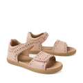 A pair of Bobux Kid+ Plus KP Logan Cameo Rose Pink Open Toe Sandals for girls, featuring light brown soles, stylish cut-out patterns, and adjustable Velcro straps over the toes and around the ankles. These leather sandals provide both comfort and style for little feet.