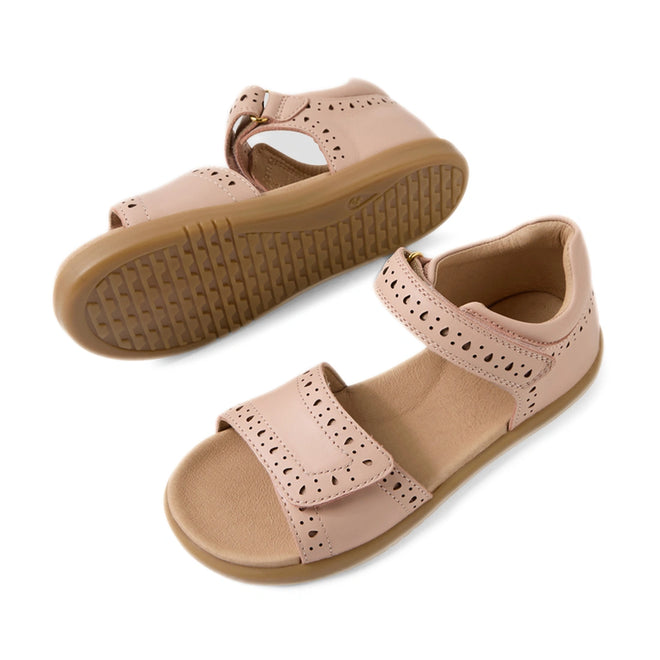 Two Bobux Kid+ Plus KP Logan Cameo Rose Pink Open Toe Sandals are displayed—one upright and one lying on its side to showcase the tan sole. Featuring decorative perforations, adjustable Velcro straps, and crafted from stylish pink leather, these sandals blend comfort with playful design for everyday adventures.