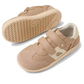 The Bobux KP Kids Plus Titan Juniors Leather Sneaker in Warm Taupe features beige leather, adjustable hook-and-loop straps, and textured slip-resistant soles. One shoe stands upright; the other is flipped to show the sole's tread pattern., 9420074855893, 9420074855909, 9420074855916, 9420074855923