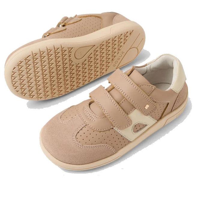 The Bobux KP Kids Plus Titan Juniors Leather Sneaker in Warm Taupe features beige leather, adjustable hook-and-loop straps, and textured slip-resistant soles. One shoe stands upright; the other is flipped to show the sole's tread pattern., 9420074855893, 9420074855909, 9420074855916, 9420074855923