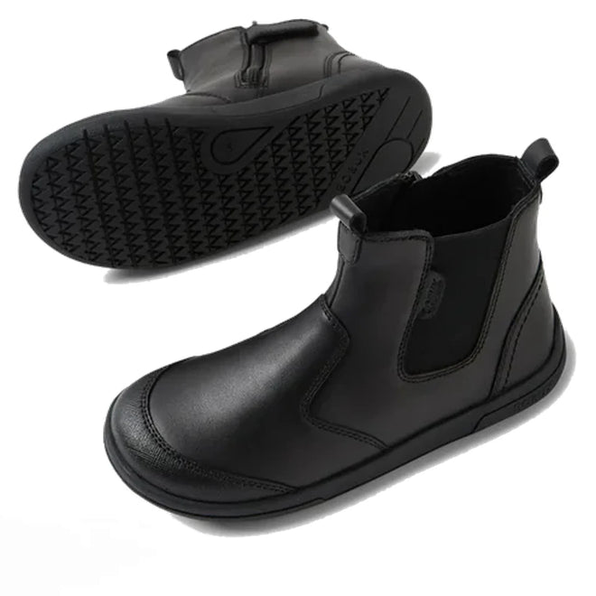 The Bobux Kid+ Black School Leather Boot Jodhpur II features black leather, ankle-high design, pull tabs, side elastic panels, and textured black rubber soles. One boot is upright; the other displays the sole’s pattern.