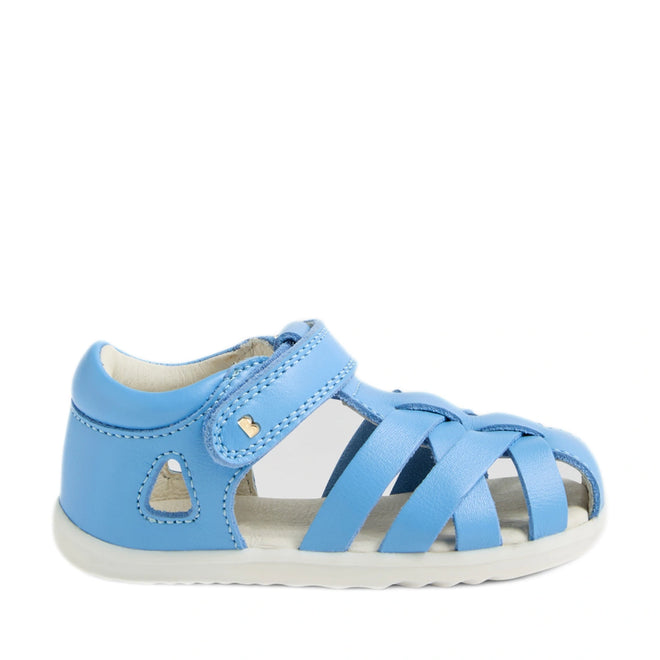 The Bobux Step Up SU Tropicana II Baltic Sea Blue Closed Toe Sandal is a blue toddler sandal inspired by the iconic Bobux Tropicana design. It features a closed heel, open toe, and crisscross straps, along with a Velcro strap and a small heart detail near the ankle. Like many toddler leather sandals, it has a white, flat sole., 9420074809957, 9420074809964, 9420074809971
